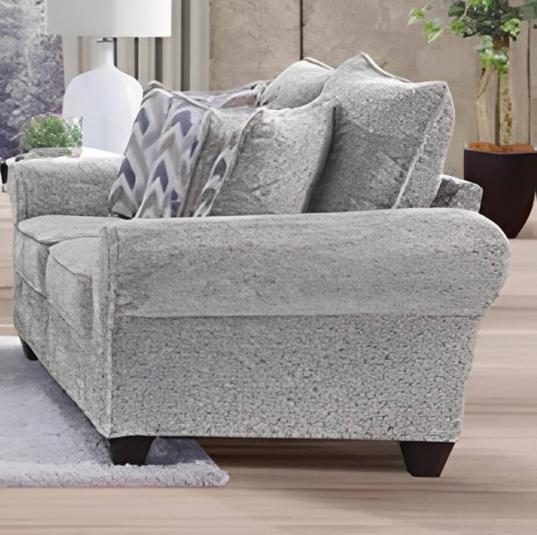 A light gray textured fabric loveseat sofa with matching throw pillows, styled in a living room setting with a lamp, rug, and potted plant.