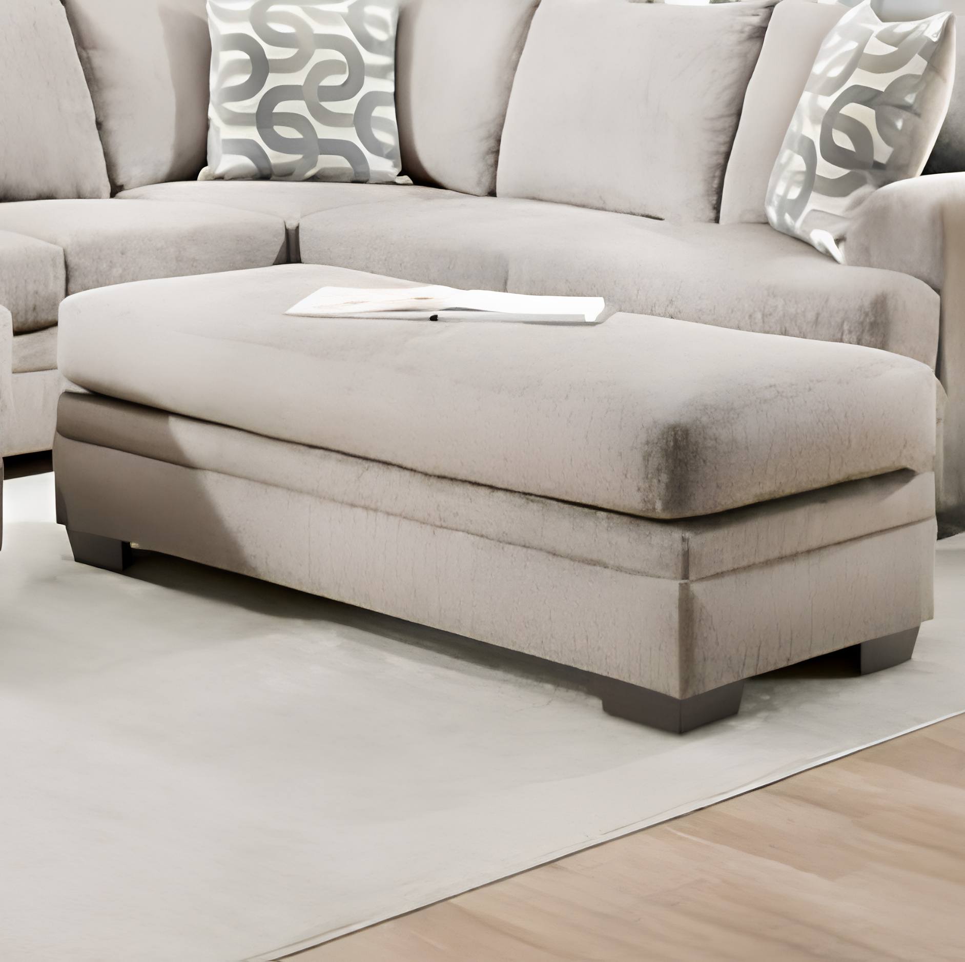 A light beige upholstered sectional sofa with a large matching ottoman in a staged living room setting, featuring patterned accent pillows and a light rug.