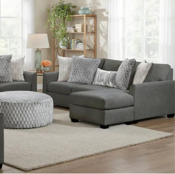 A modern gray fabric sectional sofa with a left-facing chaise lounge, styled in a bright living room with matching gray and white pillows, a round textured ottoman, and a light rug.