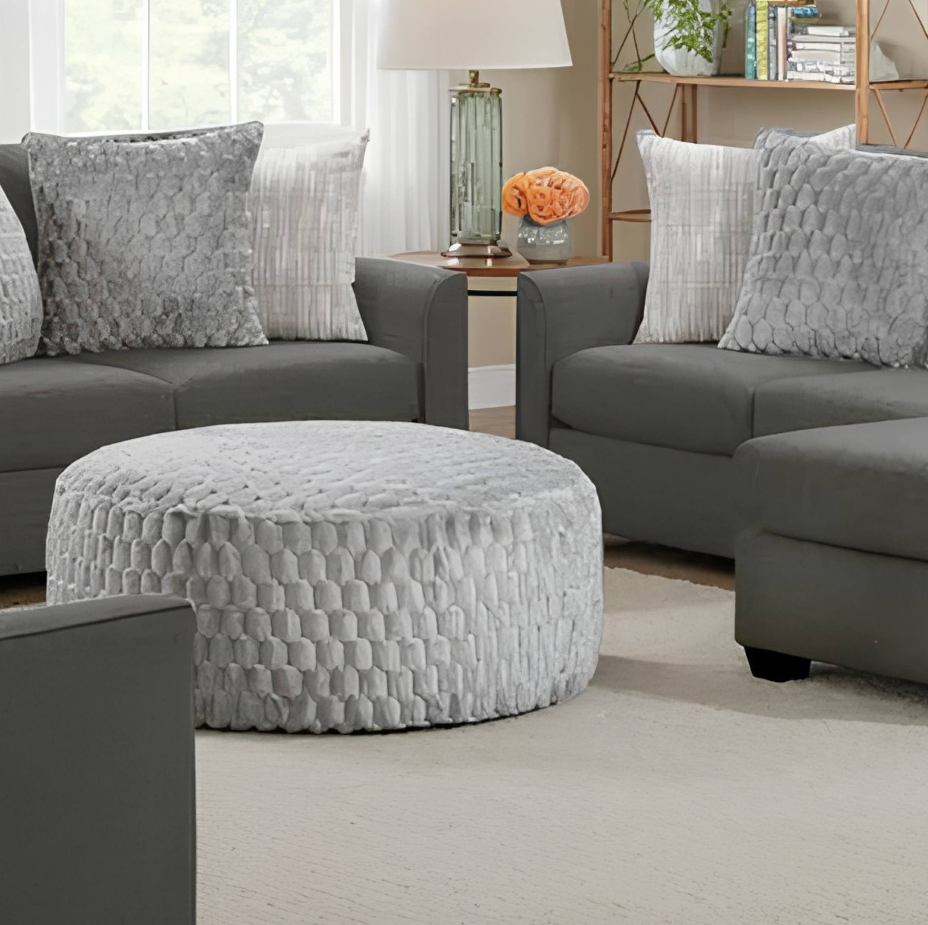 A light gray textured round pouf ottoman in a modern living room setting, styled with gray sofas and decorative pillows.