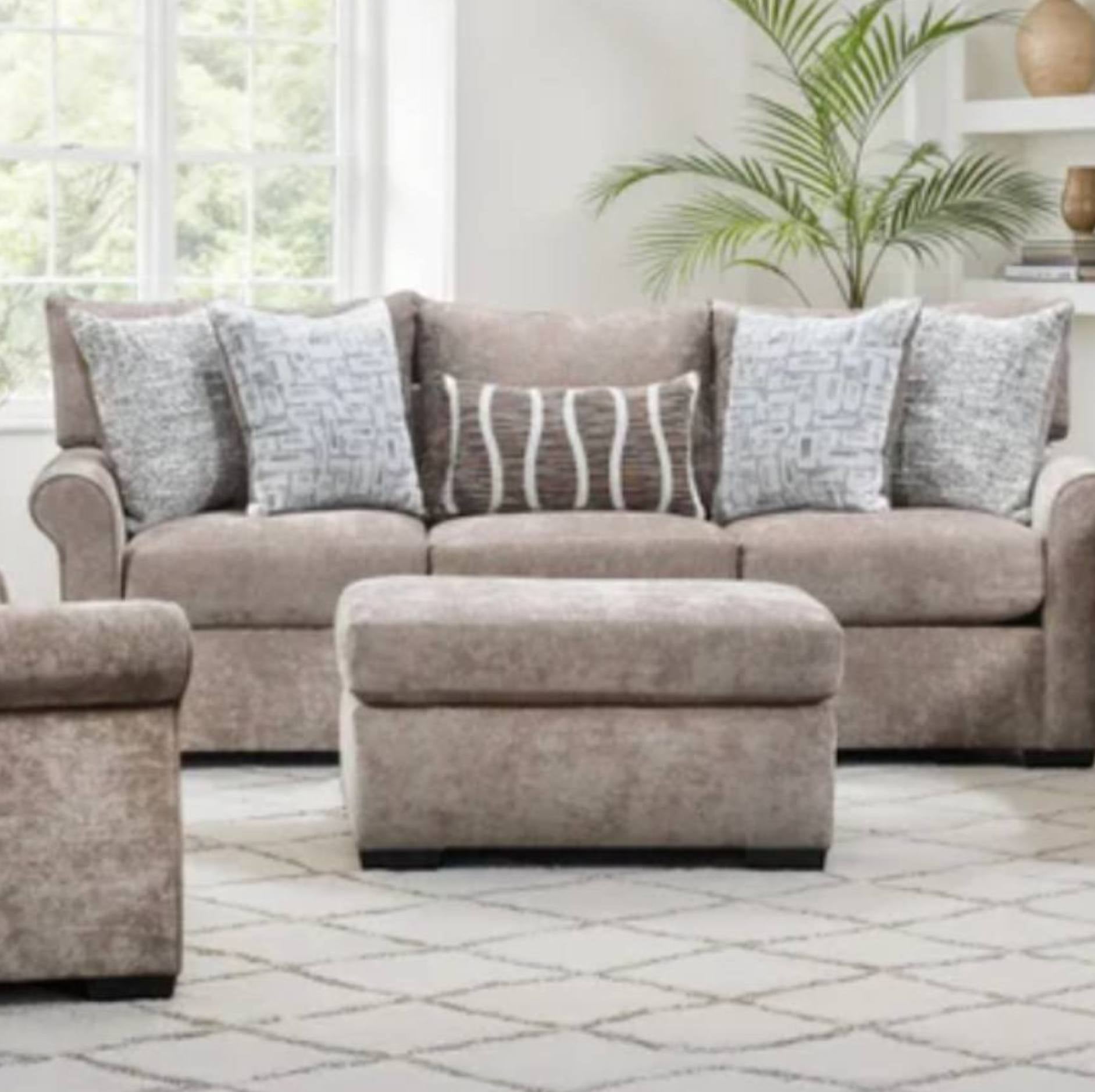 A beige upholstered sofa set in a bright living room, featuring a three-seat sofa, a matching ottoman, a partial matching armchair, and several decorative throw pillows.