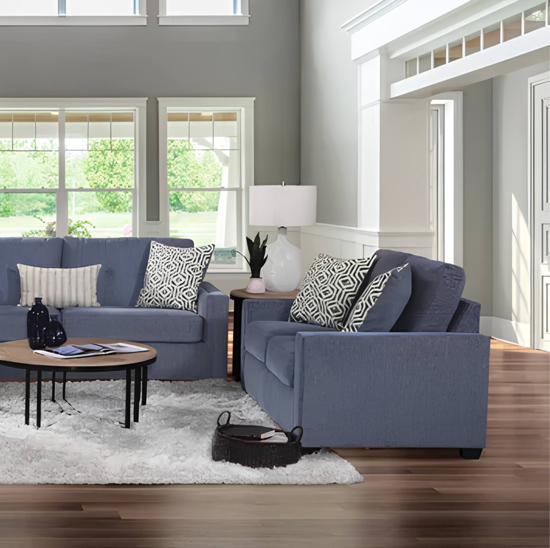 A blue fabric two-seater sofa and matching loveseat arranged in a bright living room with wood floors, a white shag rug, a round coffee table, and a white table lamp.