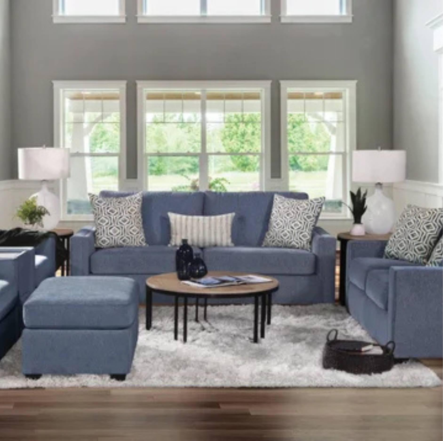 A staged living room featuring a blue fabric sofa set with patterned throw pillows, a matching blue ottoman, a round wooden coffee table, and a white shag rug in front of large windows.