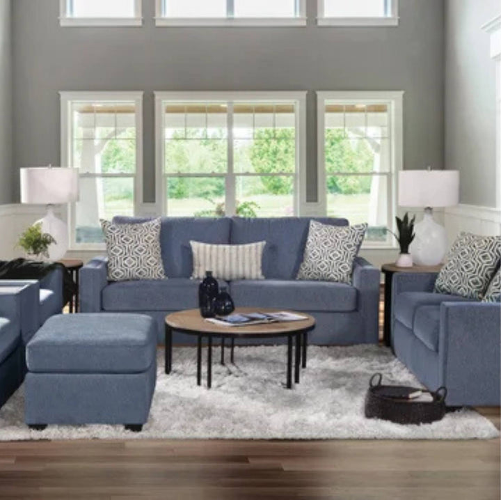 A staged living room featuring a blue fabric sofa set with patterned throw pillows, a matching blue ottoman, a round wooden coffee table, and a white shag rug in front of large windows.