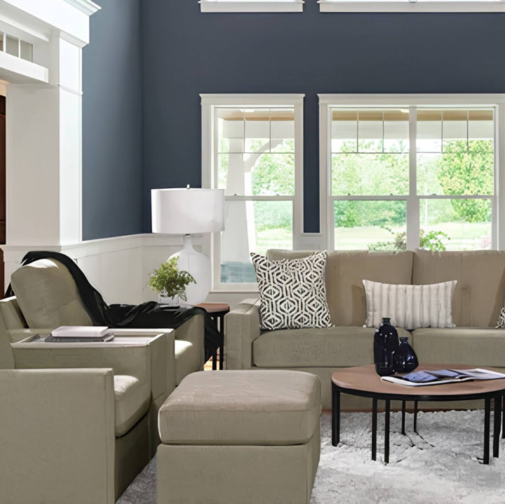 A styled living room featuring a beige sofa, matching armchair, and ottoman, decorated with patterned and striped pillows, set against a dark blue wall with white trim and windows.