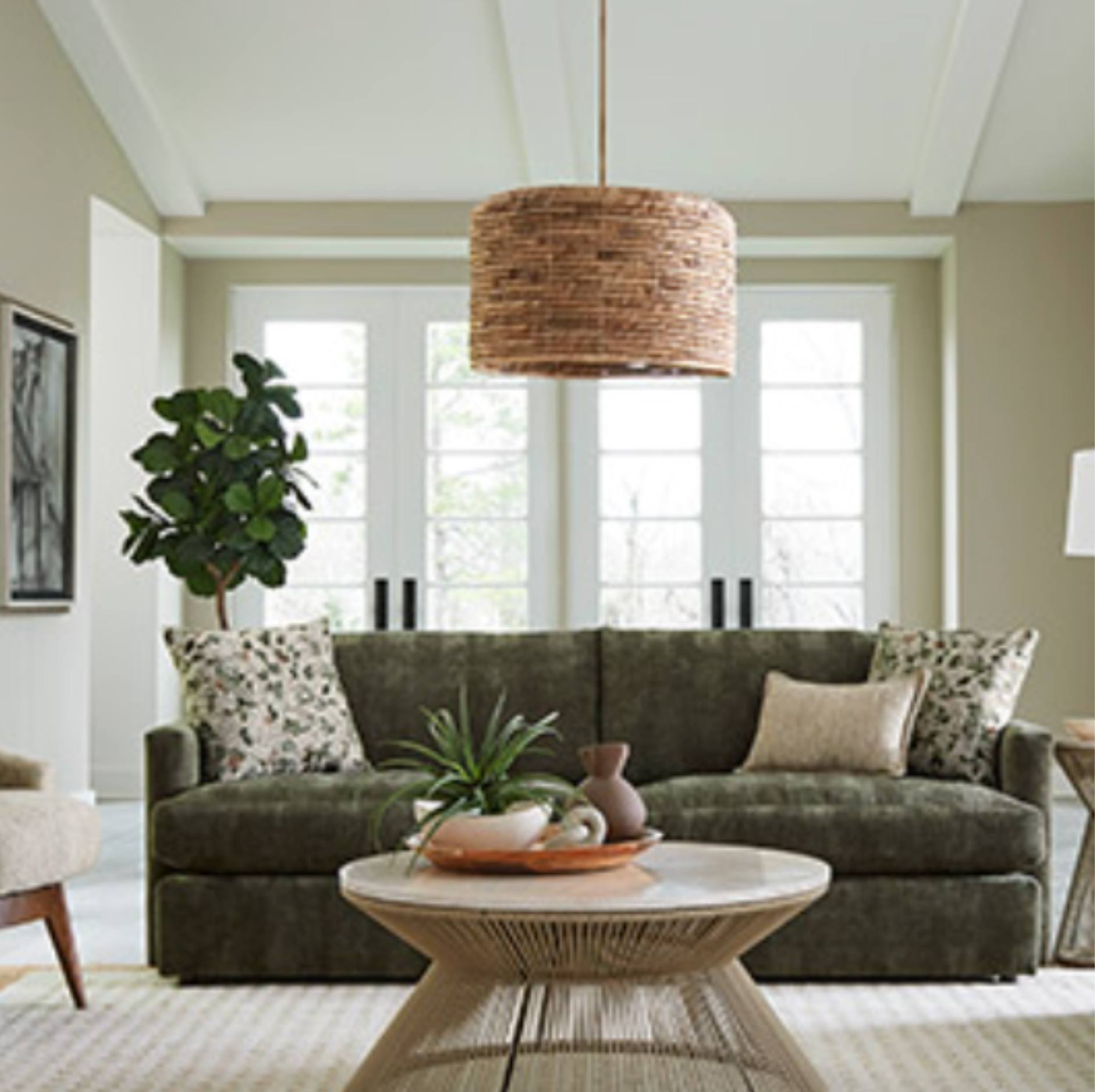 A modern living room featuring a dark olive green velvet sofa with patterned throw pillows, a round woven coffee table, and a large cylindrical wicker pendant light hanging from the ceiling.