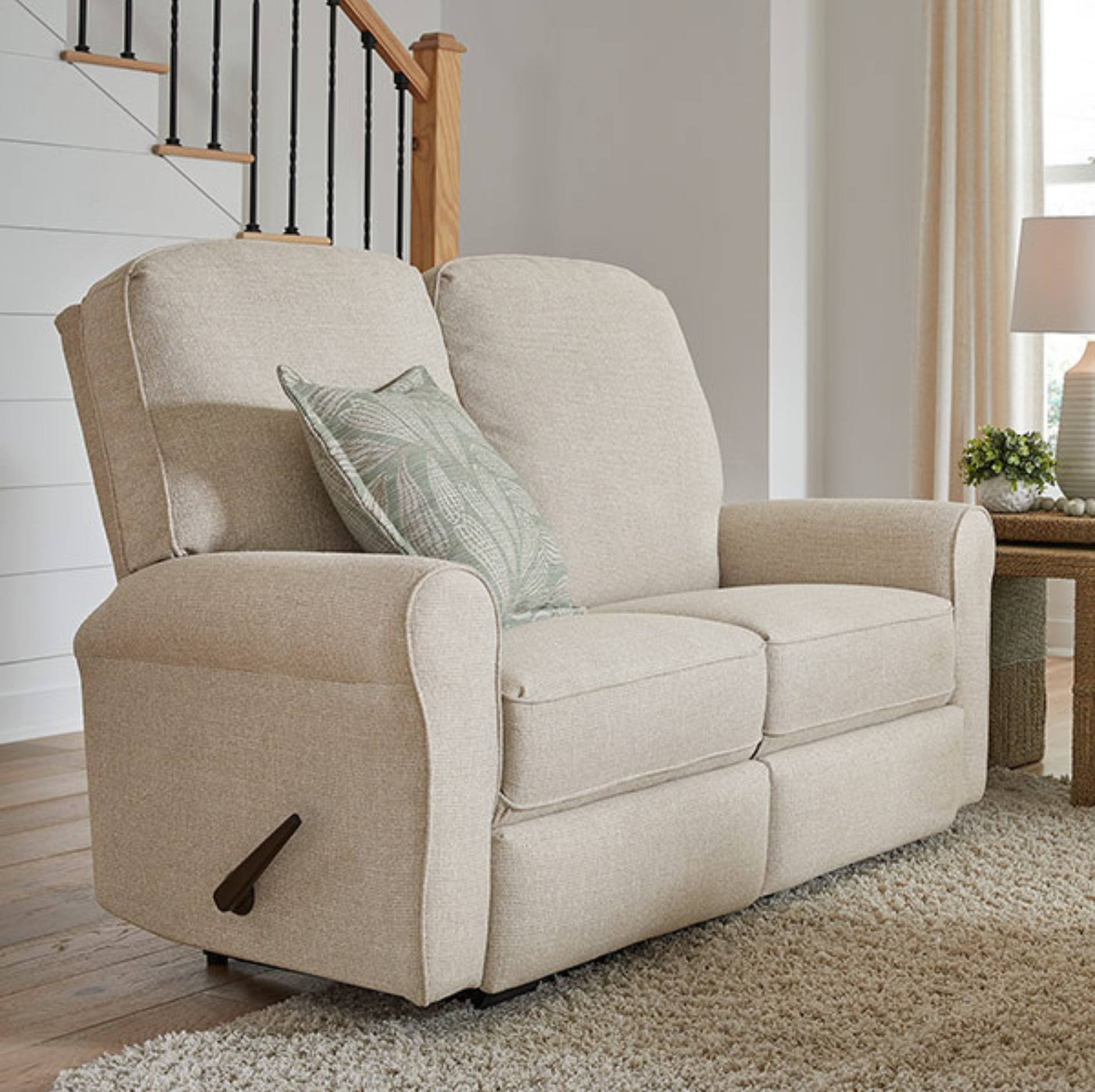 A beige upholstered recliner loveseat with a matching throw pillow, shown in a bright living room setting with a staircase, side table, and lamp.