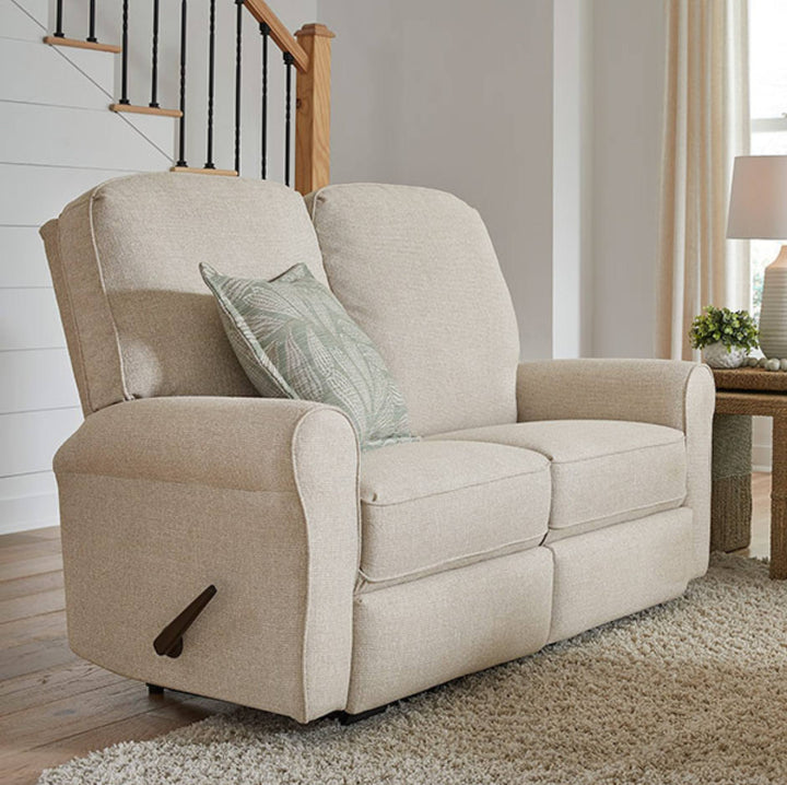A beige upholstered recliner loveseat with a matching throw pillow, shown in a bright living room setting with a staircase, side table, and lamp.