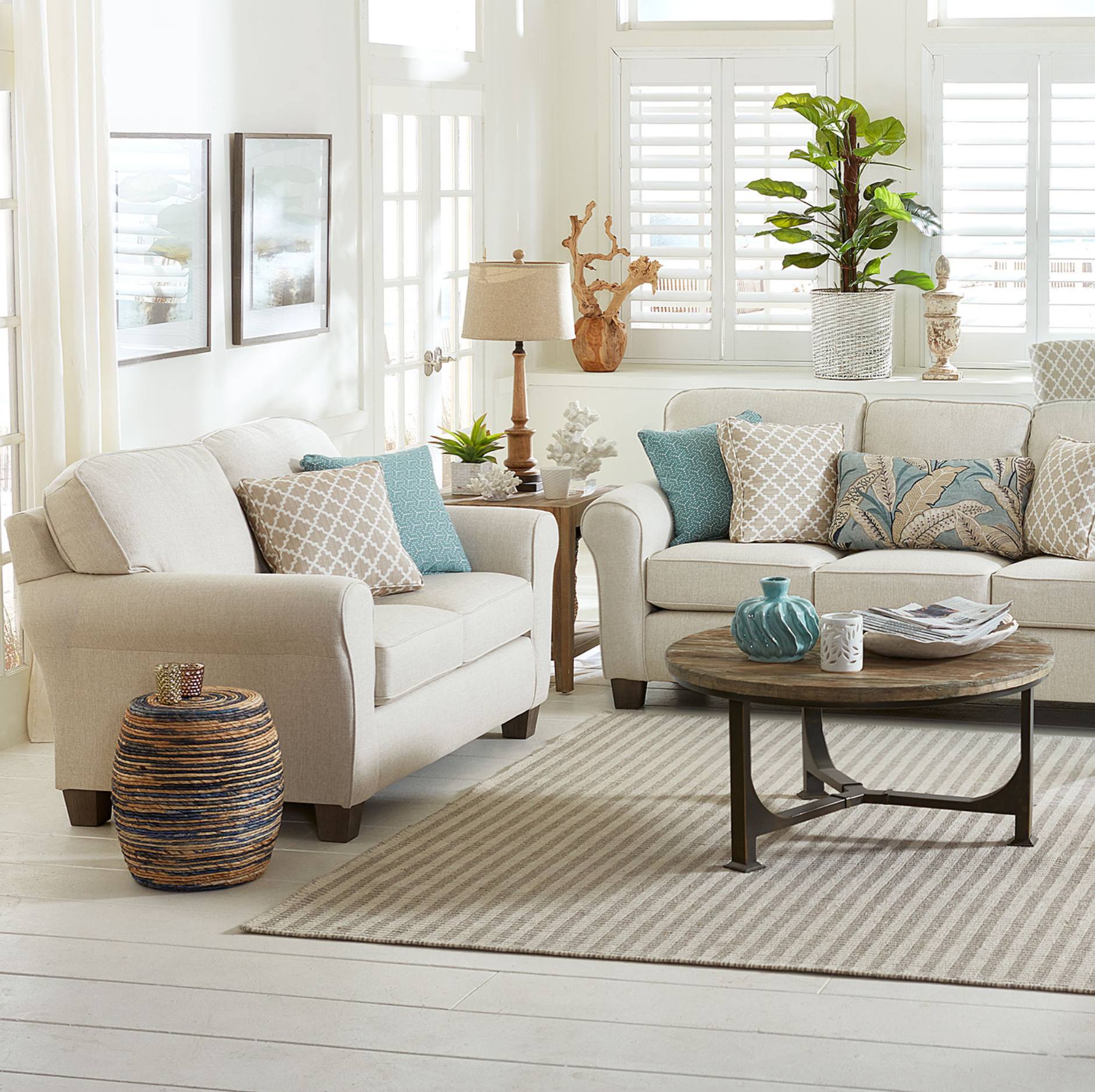 A bright, modern living room featuring a cream two-seater sofa and a matching loveseat with patterned blue and beige throw pillows, styled with a striped area rug, a round wooden coffee table, and neutral decor.