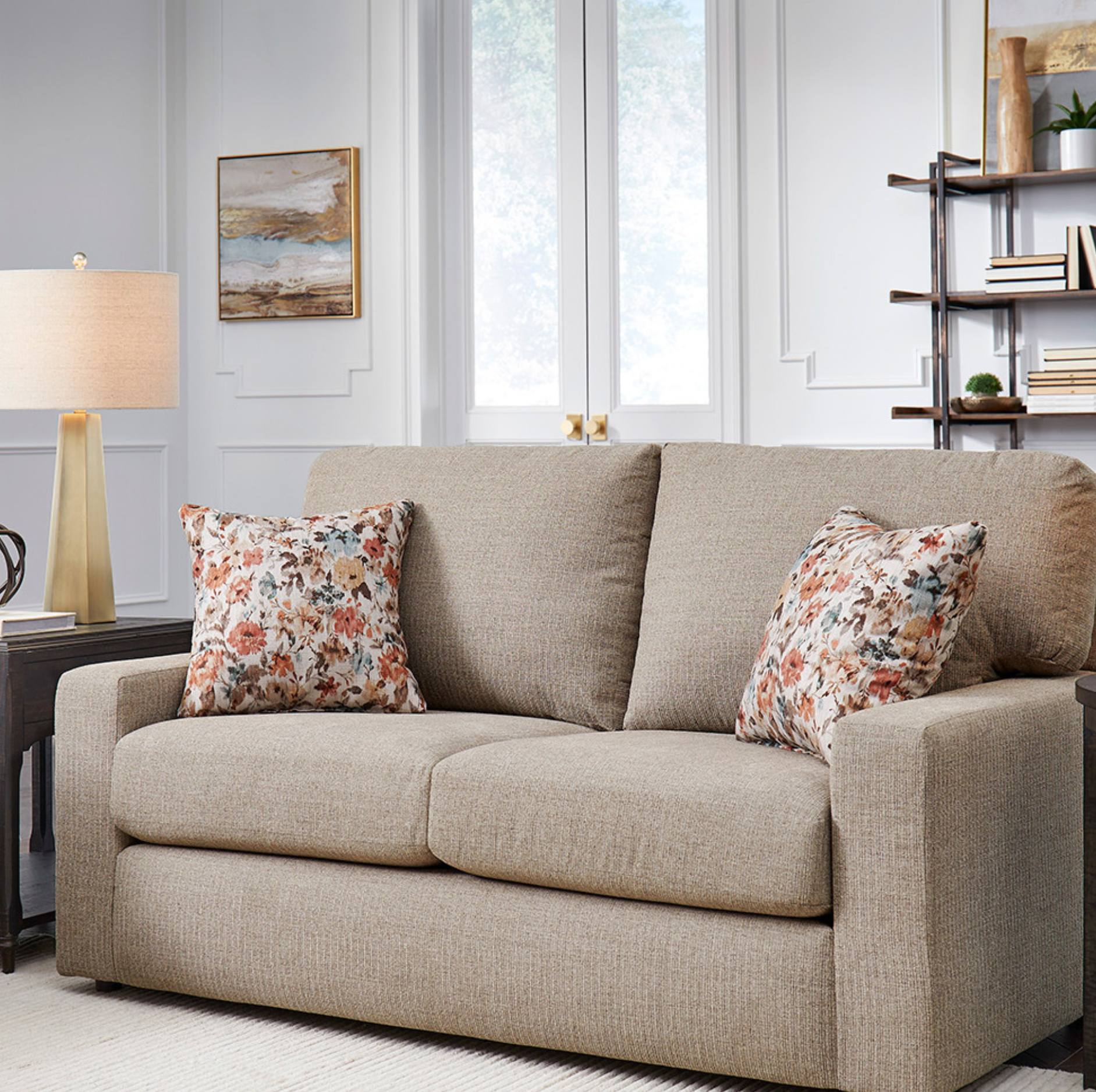 A beige upholstered loveseat in a bright living room, styled with two floral throw pillows. The sofa is positioned in front of a window, flanked by a dark wood side table with a lamp on the left and a dark shelving unit on the right.