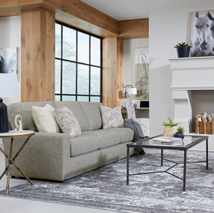 A modern living room featuring a light gray upholstered sofa as the main product, styled with patterned throw pillows, a glass-top coffee table, a gray area rug, and a rustic wood-paneled interior with a fireplace and large black-framed window.