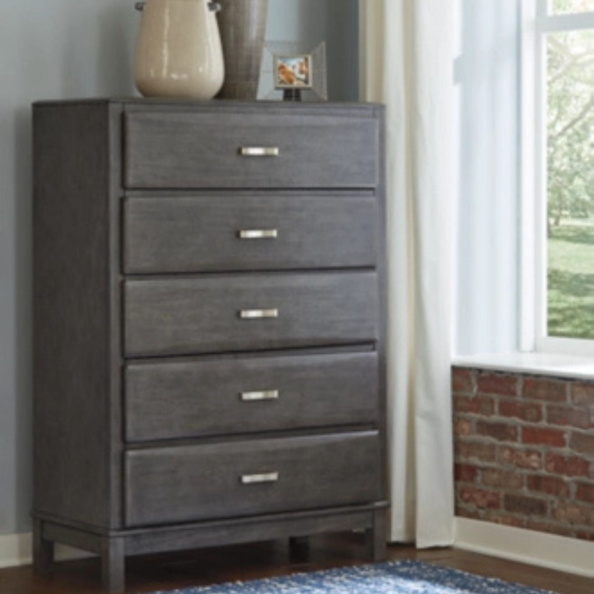 Gray dresser with five drawers in a room with a window and brick wall.
