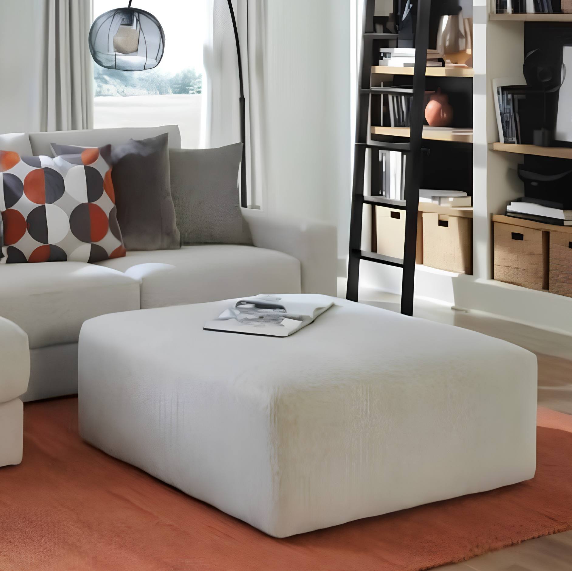 A large off-white fabric ottoman in a modern living room setting, styled with a magazine on top, next to a white sofa with patterned cushions.