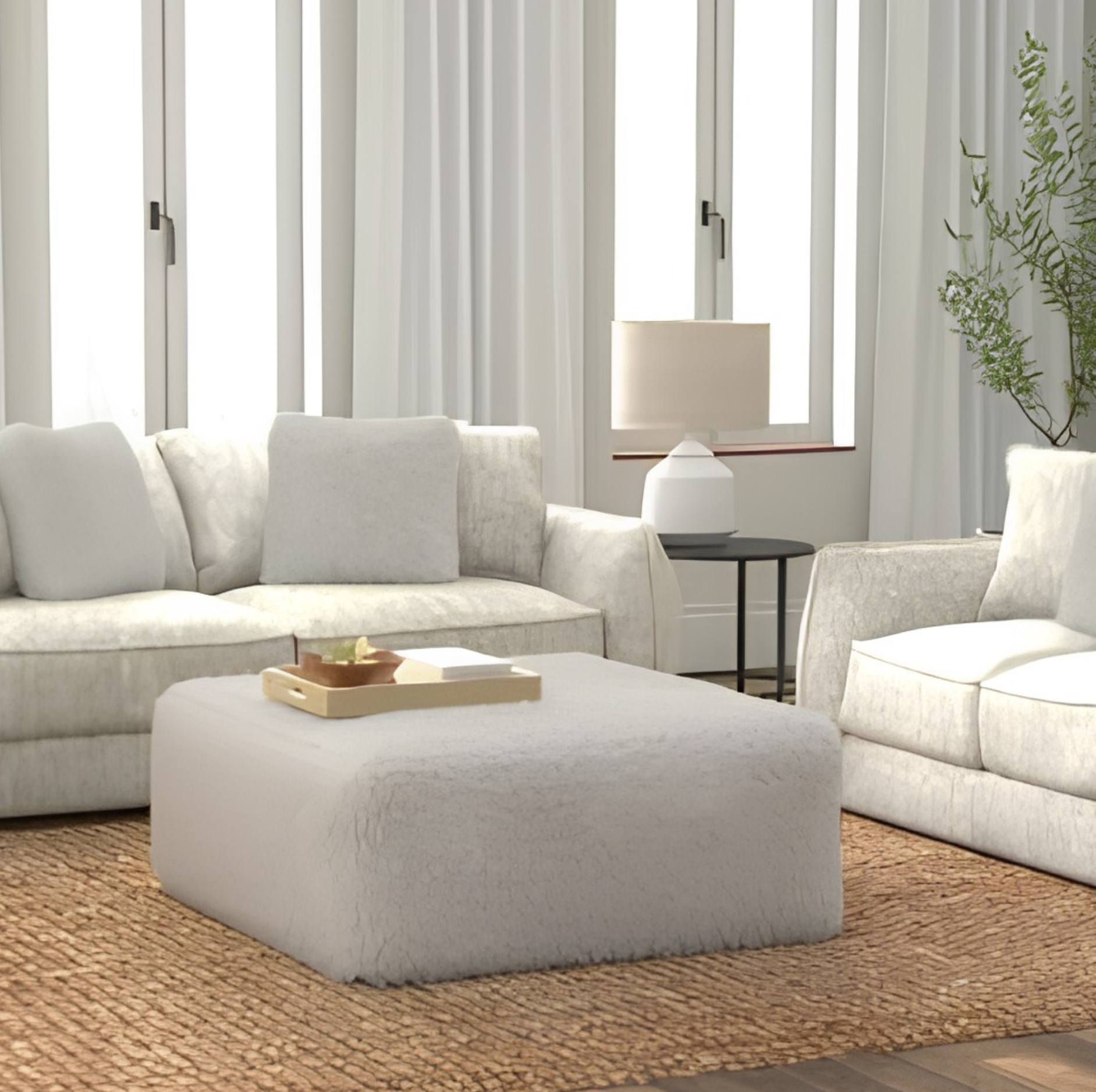 A bright, modern living room featuring a plush off-white ottoman in the foreground, a matching off-white sofa on the left, and part of a matching armchair on the right, all arranged on a woven jute rug.