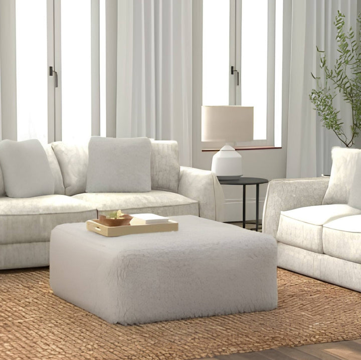 A bright, modern living room featuring a plush off-white ottoman in the foreground, a matching off-white sofa on the left, and part of a matching armchair on the right, all arranged on a woven jute rug.