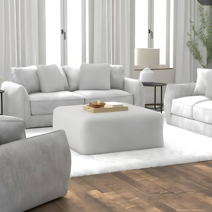 A bright, modern living room featuring a white three-seater sofa, a matching armchair, a white ottoman coffee table, and a light gray armchair on a white rug over a wood floor.