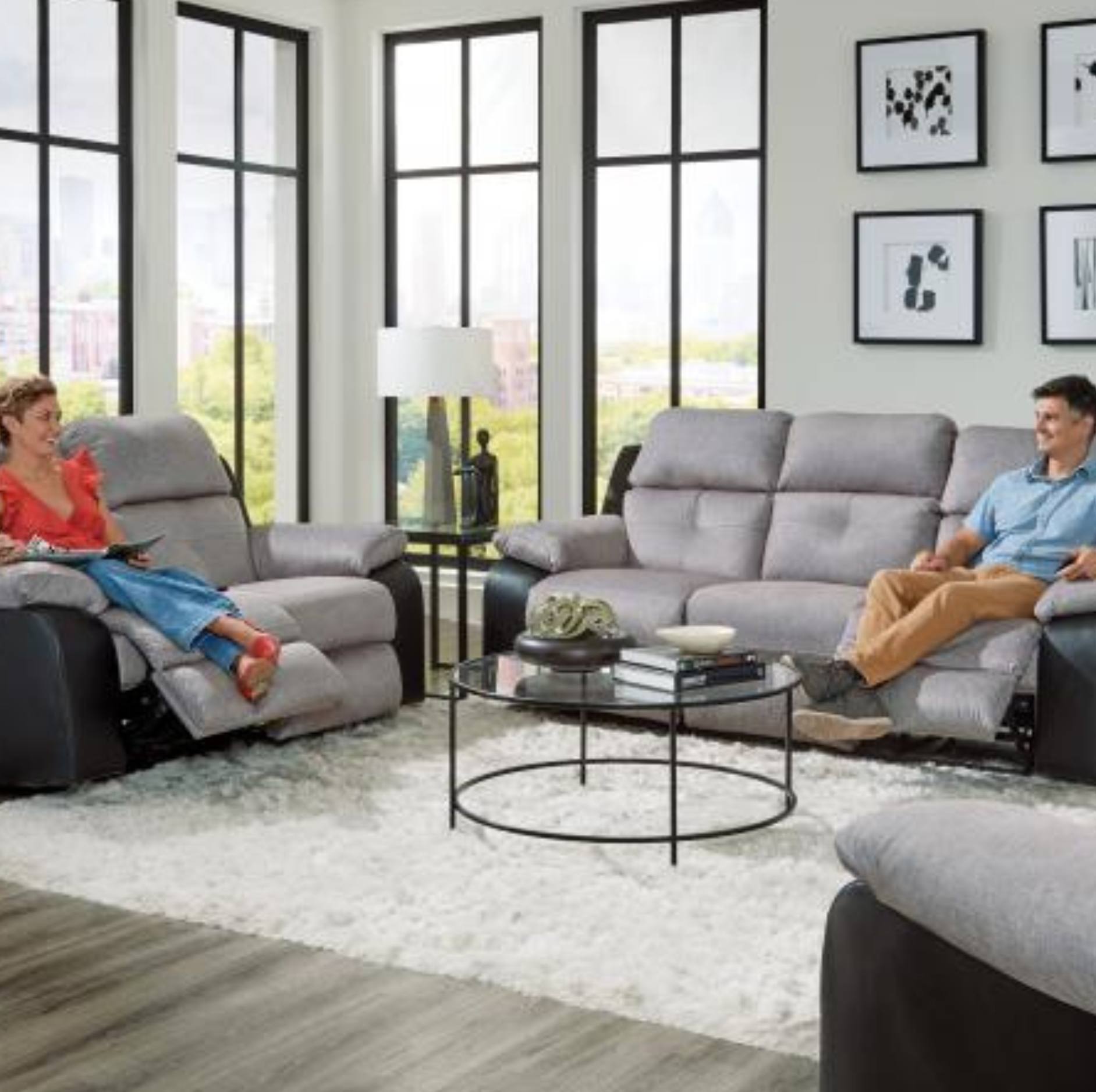 A modern living room scene showing a gray upholstered reclining loveseat with a woman sitting in it, alongside a matching gray loveseat with a man sitting in it. The room has large black-framed windows, a white shag rug, and a round glass-top coffee table.