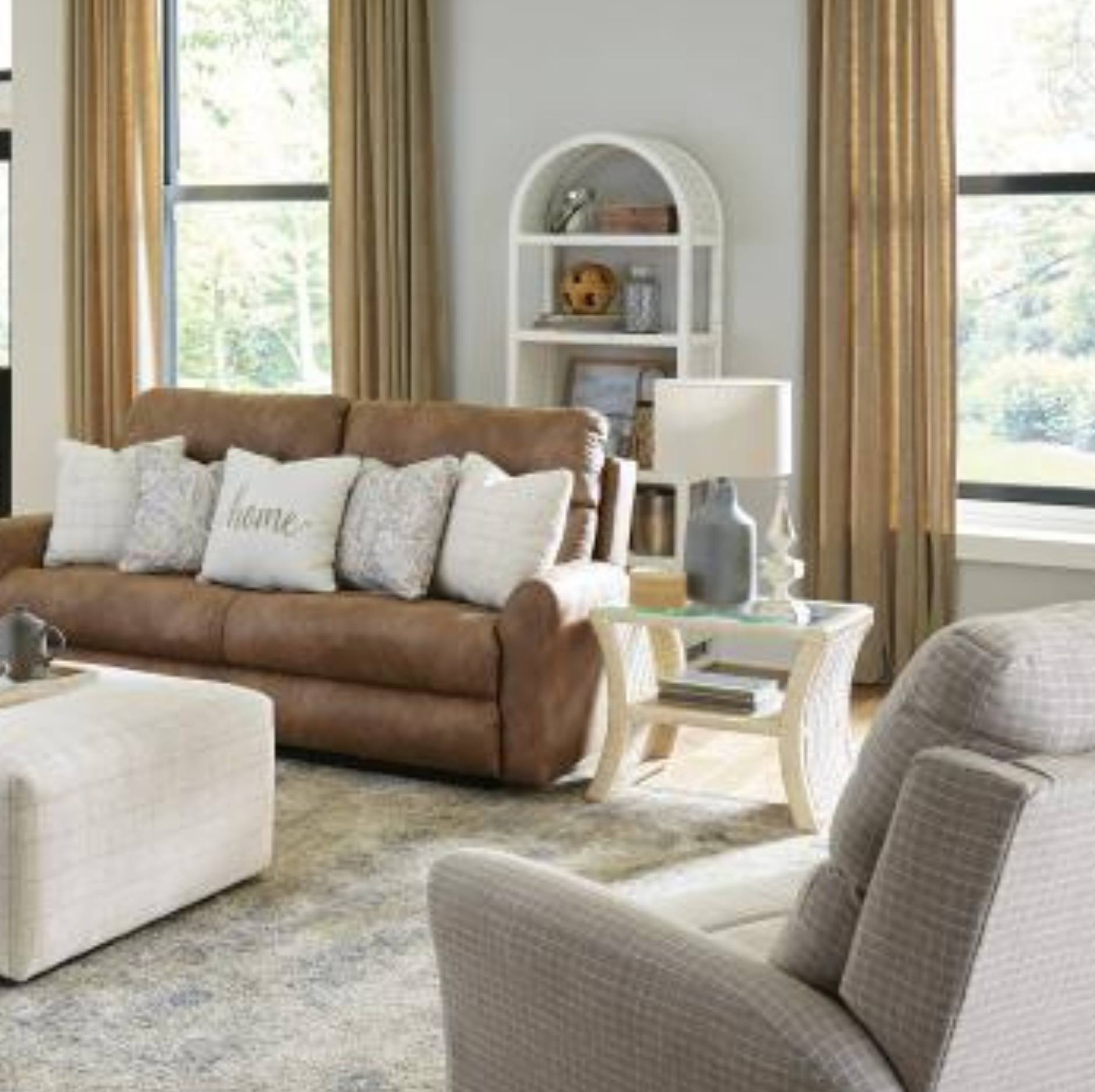 A brown leather reclining sofa in a bright, modern living room setting, styled with white and gray throw pillows, next to a matching beige armchair and an ottoman.