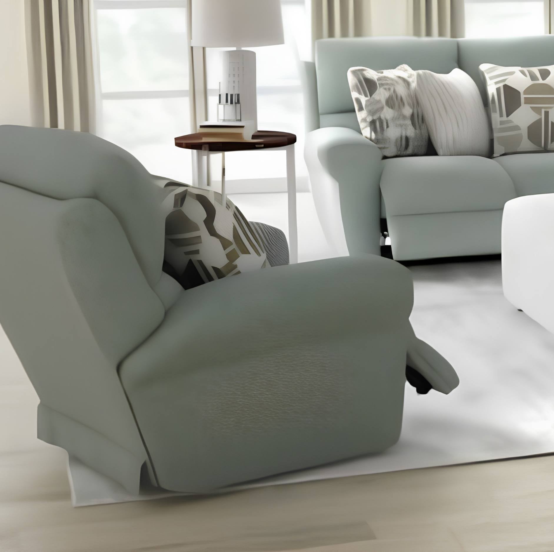 A light sage green recliner chair in a bright, modern living room setting, shown with a matching patterned throw pillow and a matching light green sofa in the background.