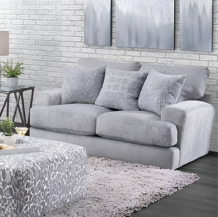 A light gray upholstered loveseat with three matching throw pillows, shown in a styled living room setting with a patterned rug, side table, and brick wall background.