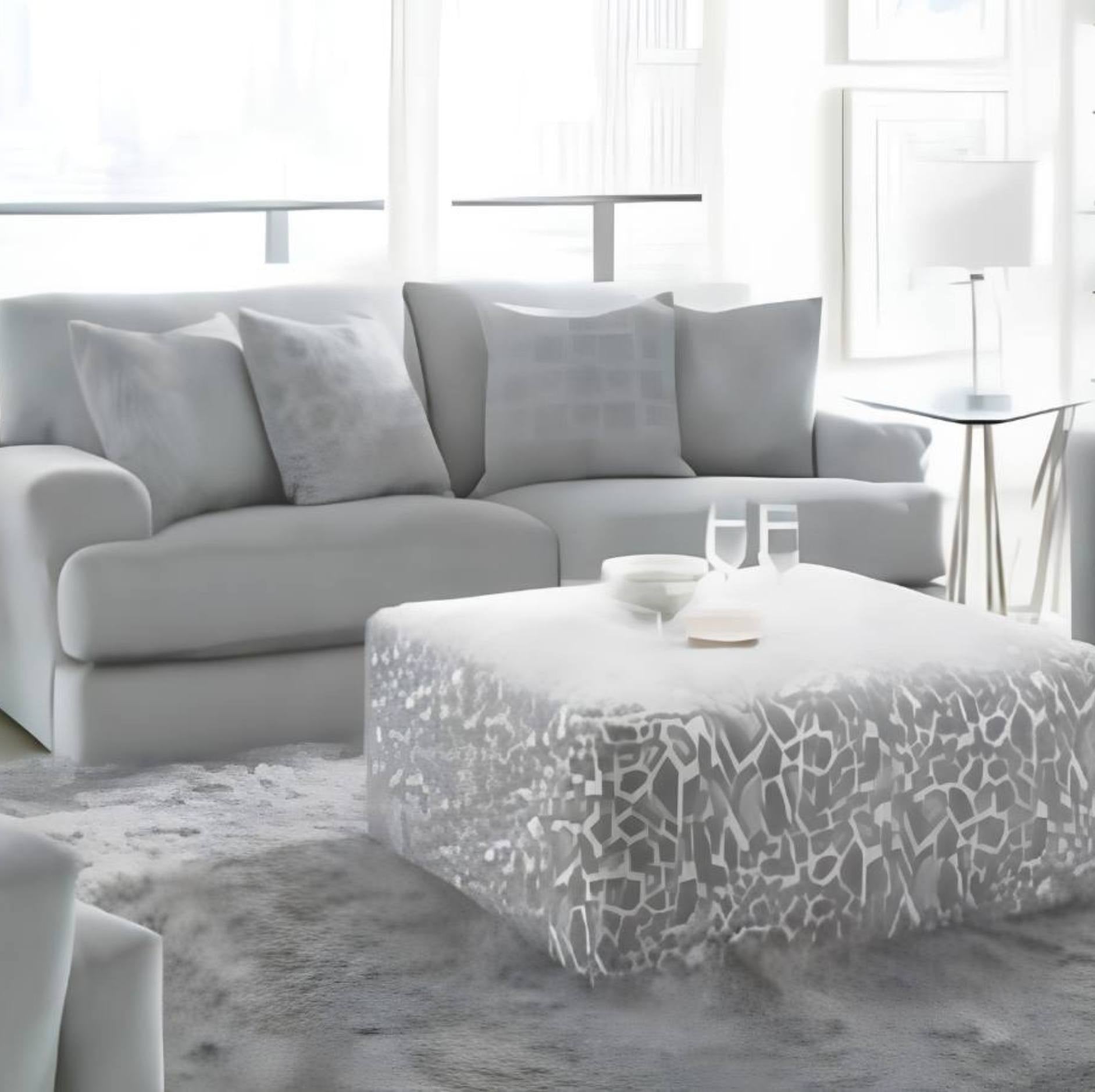 A modern living room scene featuring a light gray sectional sofa with matching throw pillows, a white ottoman with a gray abstract pattern, and a gray shag rug in a bright, airy interior.