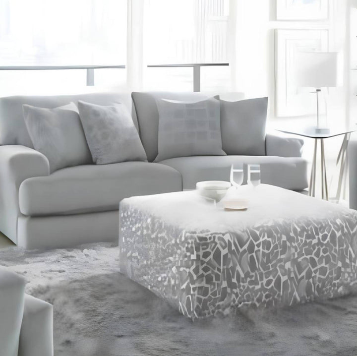 A modern living room scene featuring a light gray sectional sofa with matching throw pillows, a white ottoman with a gray abstract pattern, and a gray shag rug in a bright, airy interior.