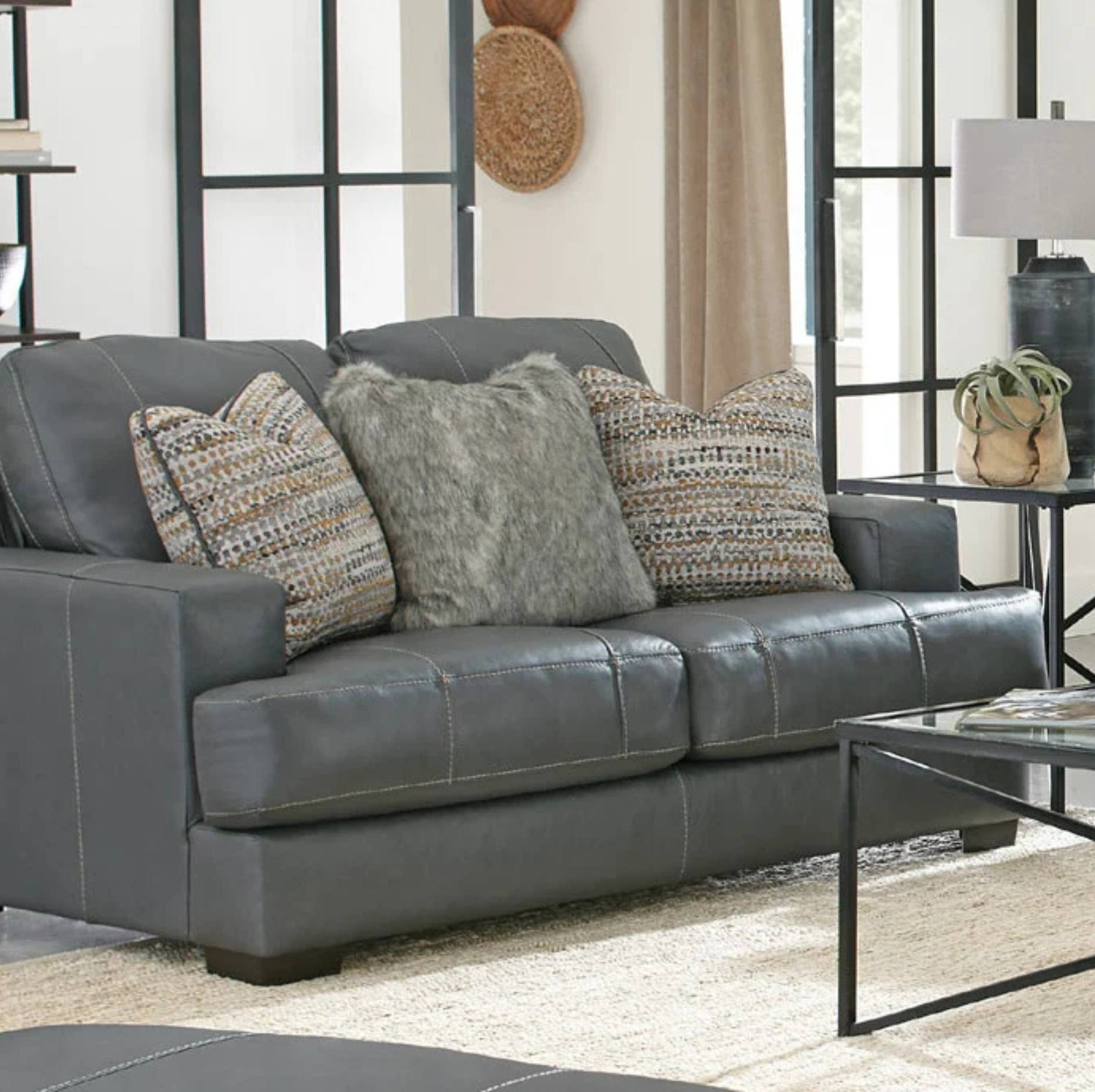 A dark gray leather loveseat sofa in a modern living room setting, styled with four decorative throw pillows including two textured gray faux fur pillows and two patterned beige and gray pillows.