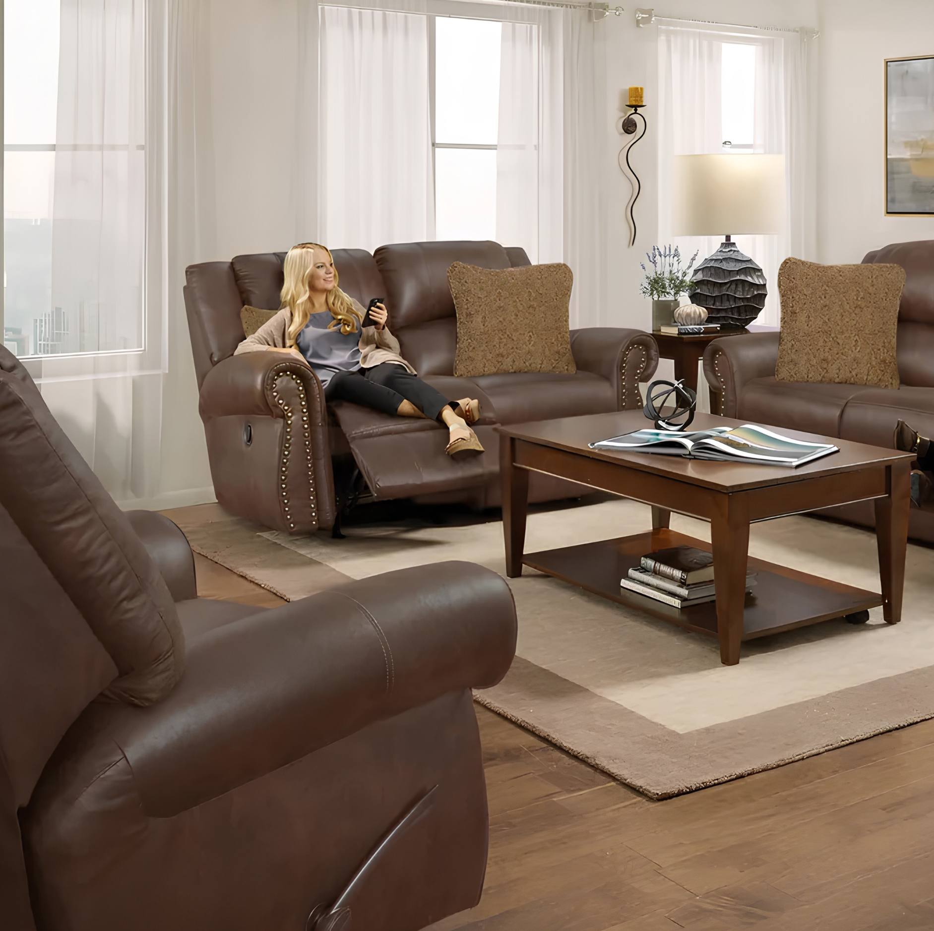 A woman relaxes in a brown leather recliner sofa in a bright living room. The room features a matching brown leather coffee table, a second brown leather recliner in the foreground, and a third recliner on the right.