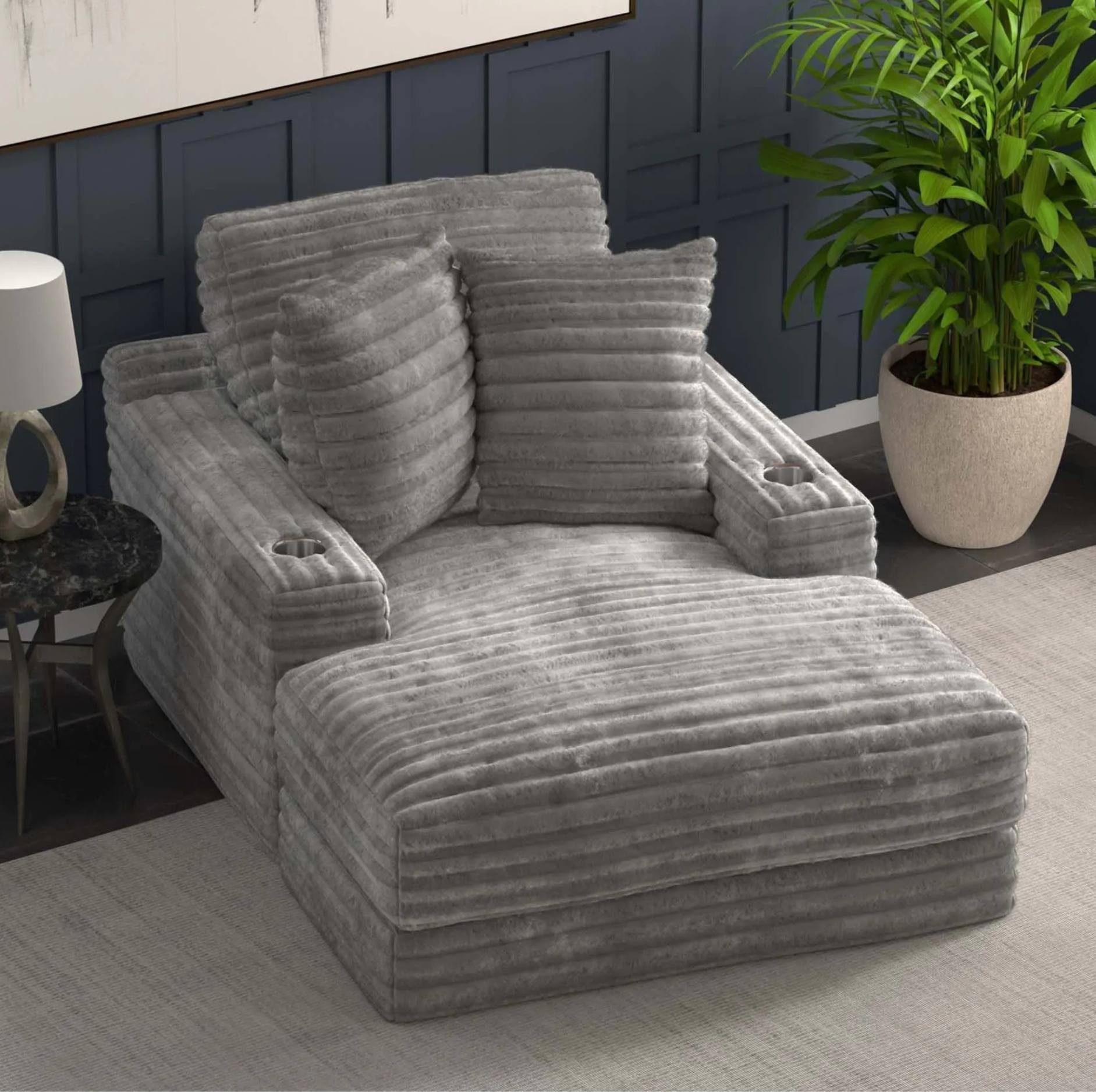 A gray ribbed chaise lounge chair with two matching throw pillows, shown in a styled living room setting with a dark paneled wall, a potted plant, and a side table.
