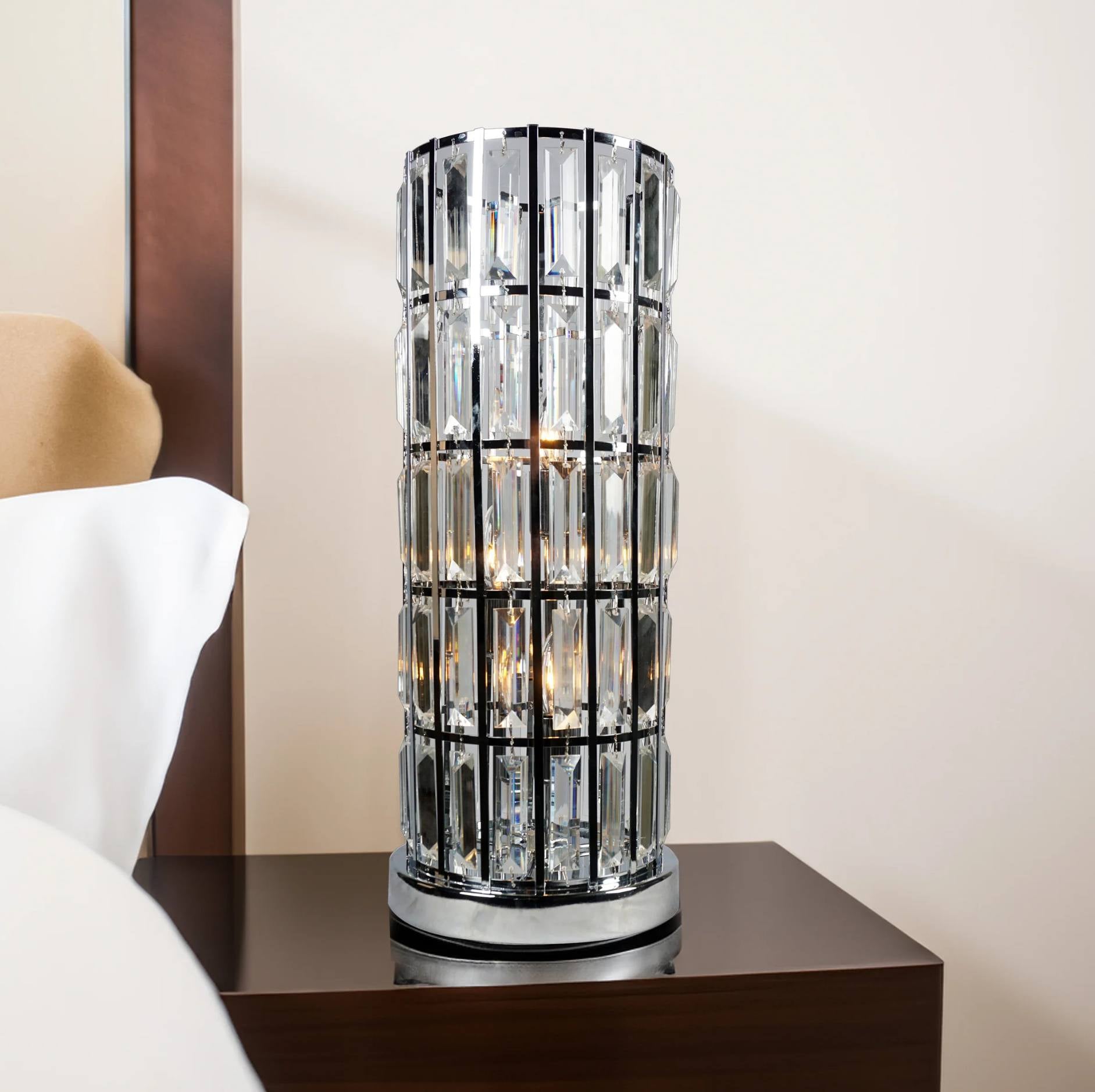 A modern cylindrical table lamp with a clear faceted crystal or glass column and a silver-toned metal frame, shown on a dark wood bedside table in a bedroom setting.