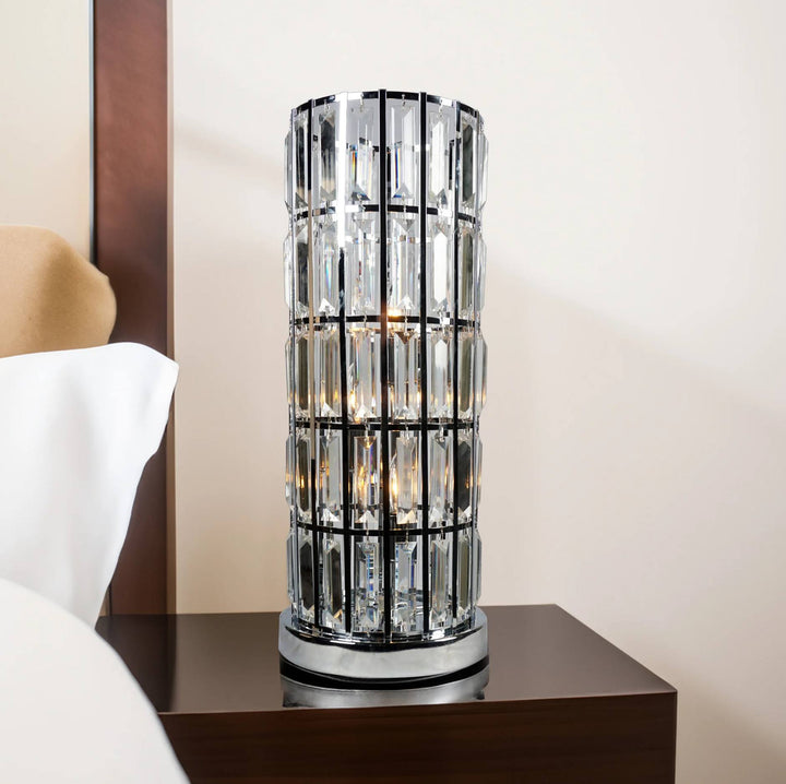 A modern cylindrical table lamp with a clear faceted crystal or glass column and a silver-toned metal frame, shown on a dark wood bedside table in a bedroom setting.