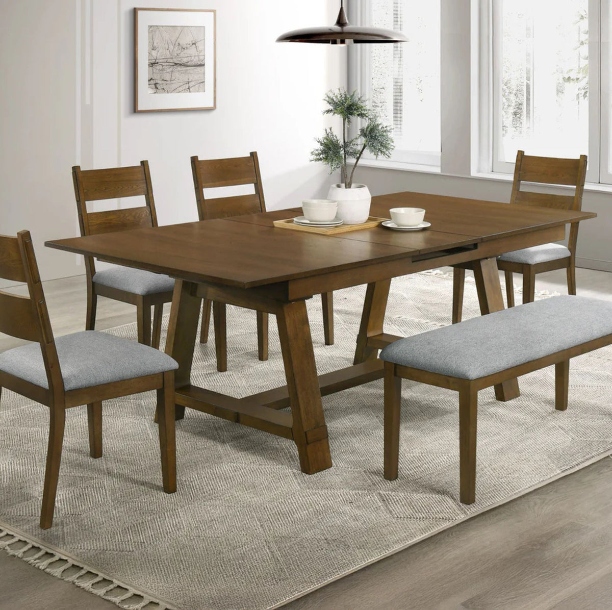 A 5-piece wooden dining room set including a rectangular table, four chairs with light gray upholstered seats, and a matching bench with a cushion, arranged on a textured rug in a bright dining room.
