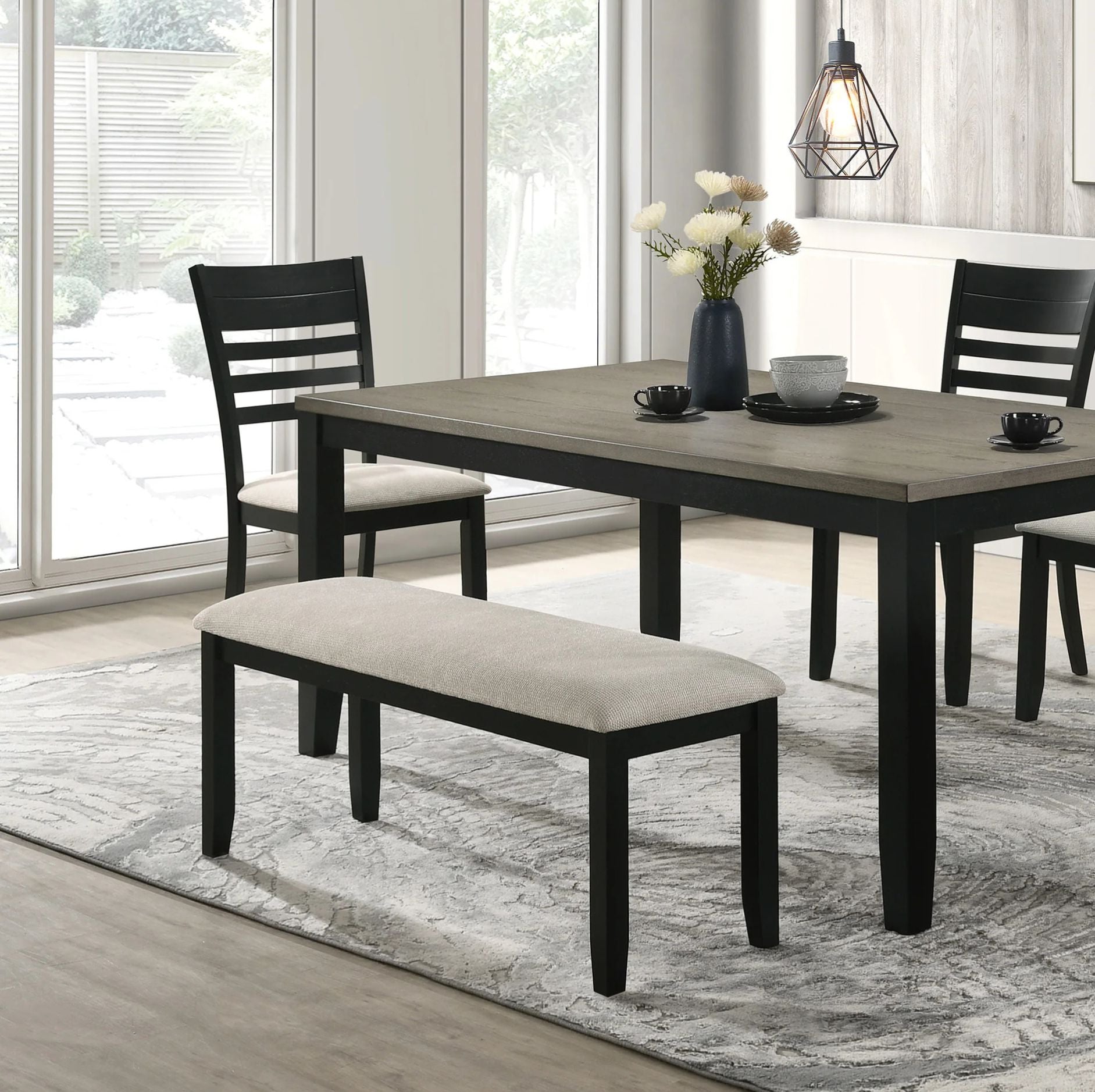 A 5-piece dining set featuring a rectangular table with a gray wood-grain top and black frame, accompanied by two black chairs with cream upholstered seats and a matching bench, set on a gray patterned rug in a bright dining room.