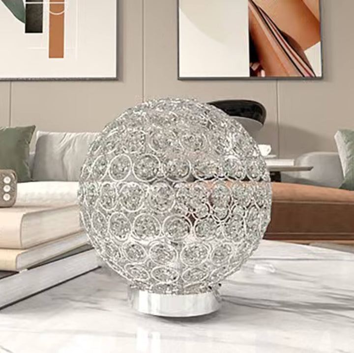 A spherical crystal table lamp with a clear faceted glass sphere and a shiny silver base, placed on a marble tabletop in a modern living room setting.