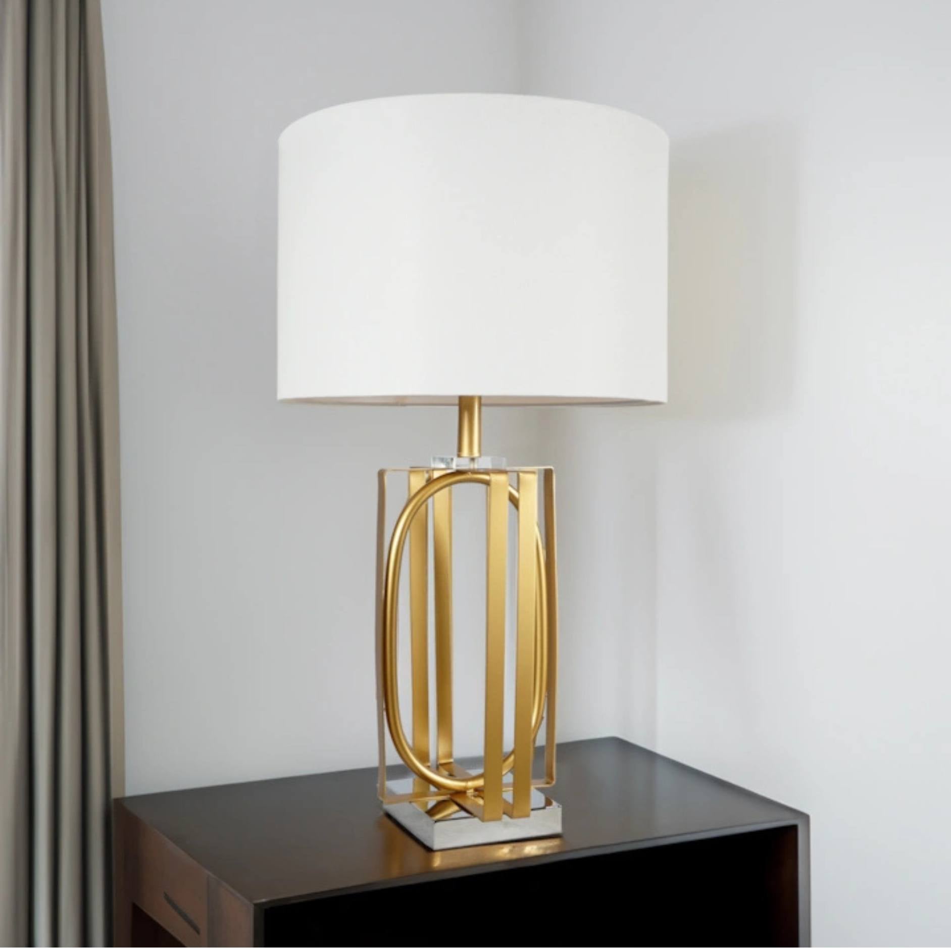 A modern table lamp with a white drum shade and a gold-finished geometric metal base, displayed on a dark wooden side table against a light gray wall.