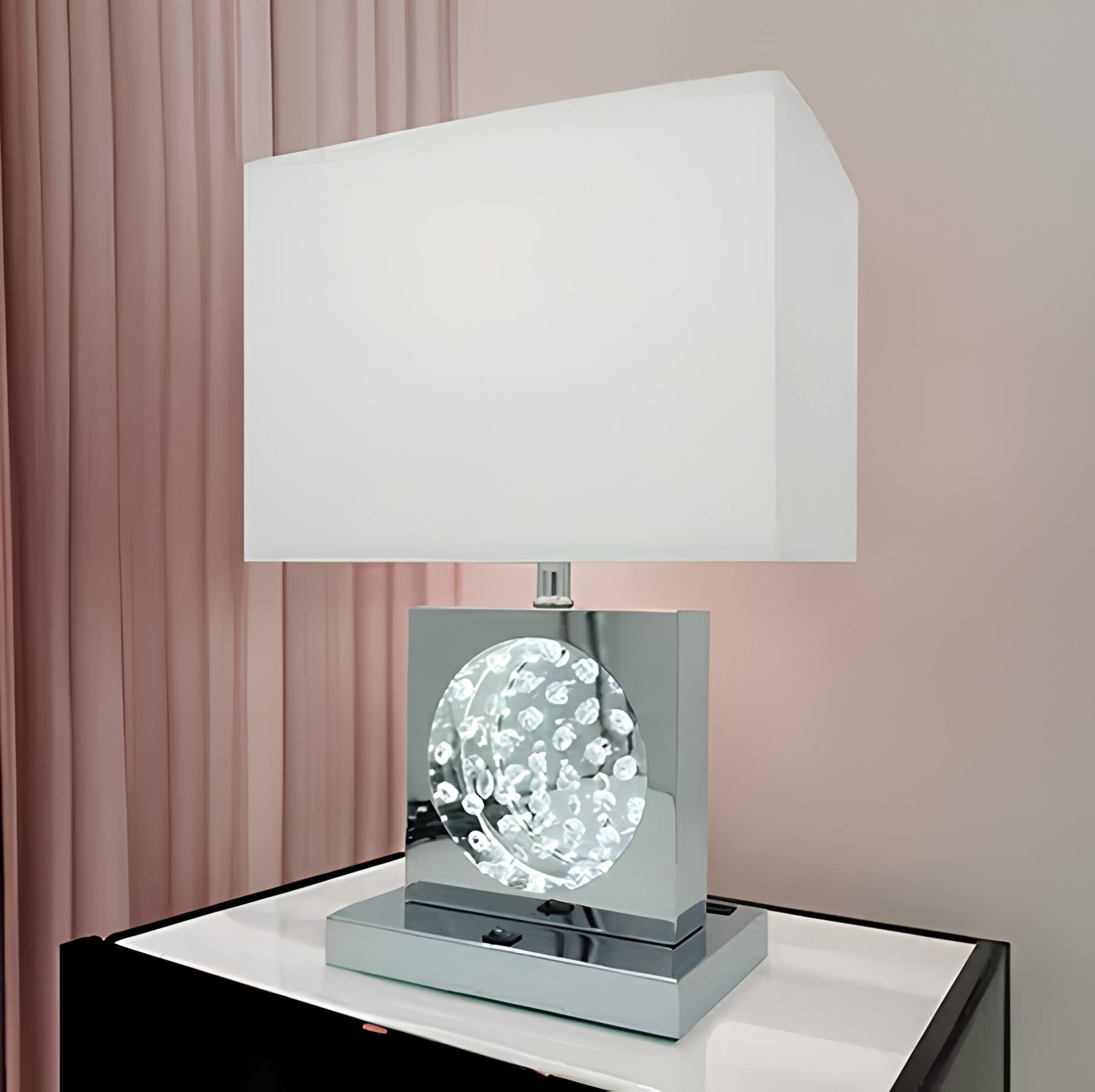 A modern table lamp with a white rectangular fabric shade, a polished silver metallic base, and a clear faceted crystal globe ornament in the center, displayed on a white surface against a soft pink curtain background.