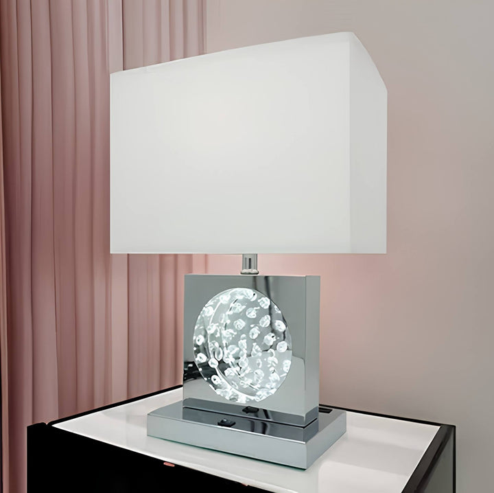 A modern table lamp with a white rectangular fabric shade, a polished silver metallic base, and a clear faceted crystal globe ornament in the center, displayed on a white surface against a soft pink curtain background.