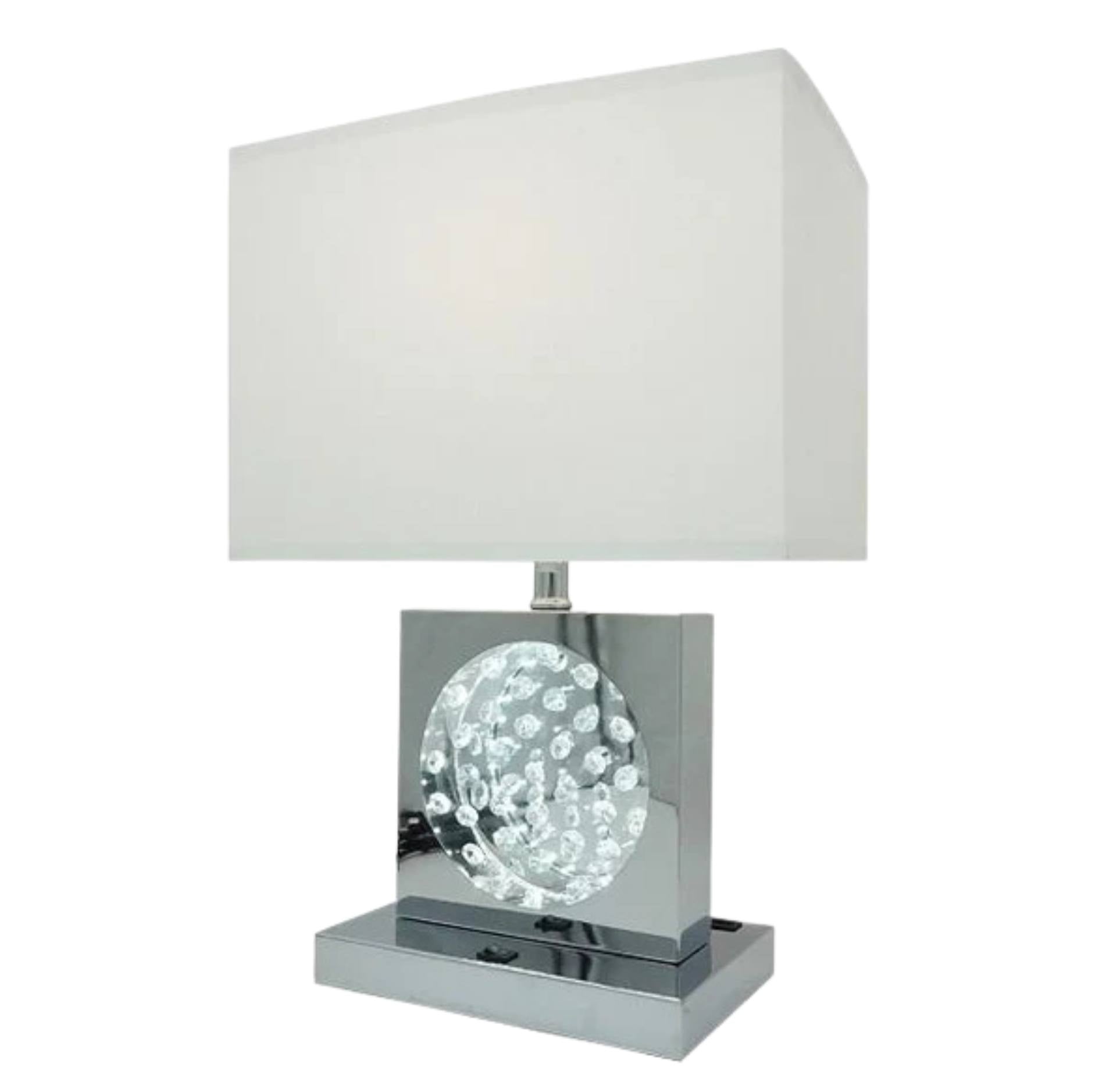 A modern table lamp with a rectangular white fabric shade, a polished chrome square base, and a clear faceted crystal sphere in the center.