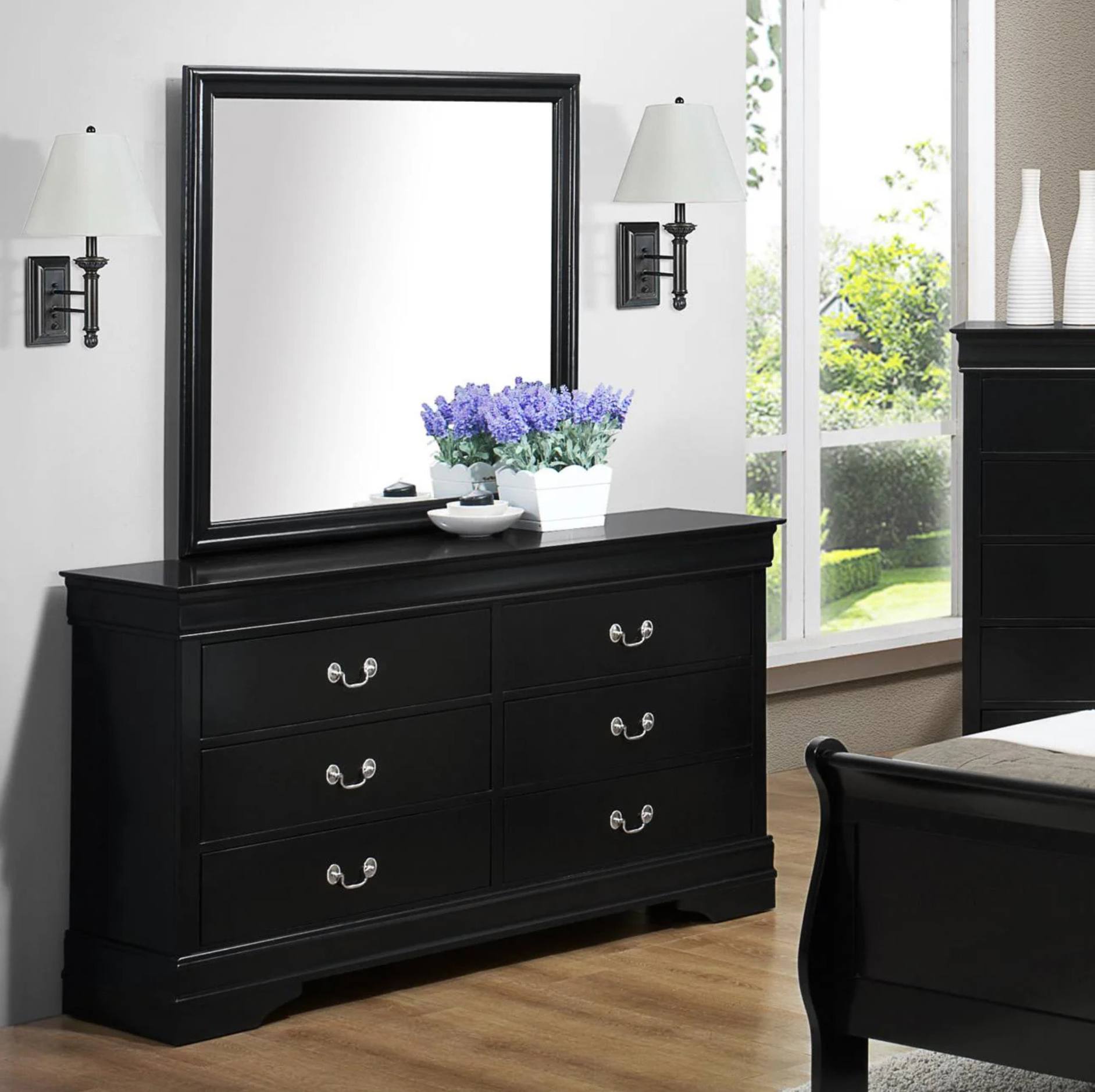 A black wooden dresser with six drawers and a matching rectangular mirror, styled in a bedroom setting with wall sconces, a window, and a bed frame partially visible.