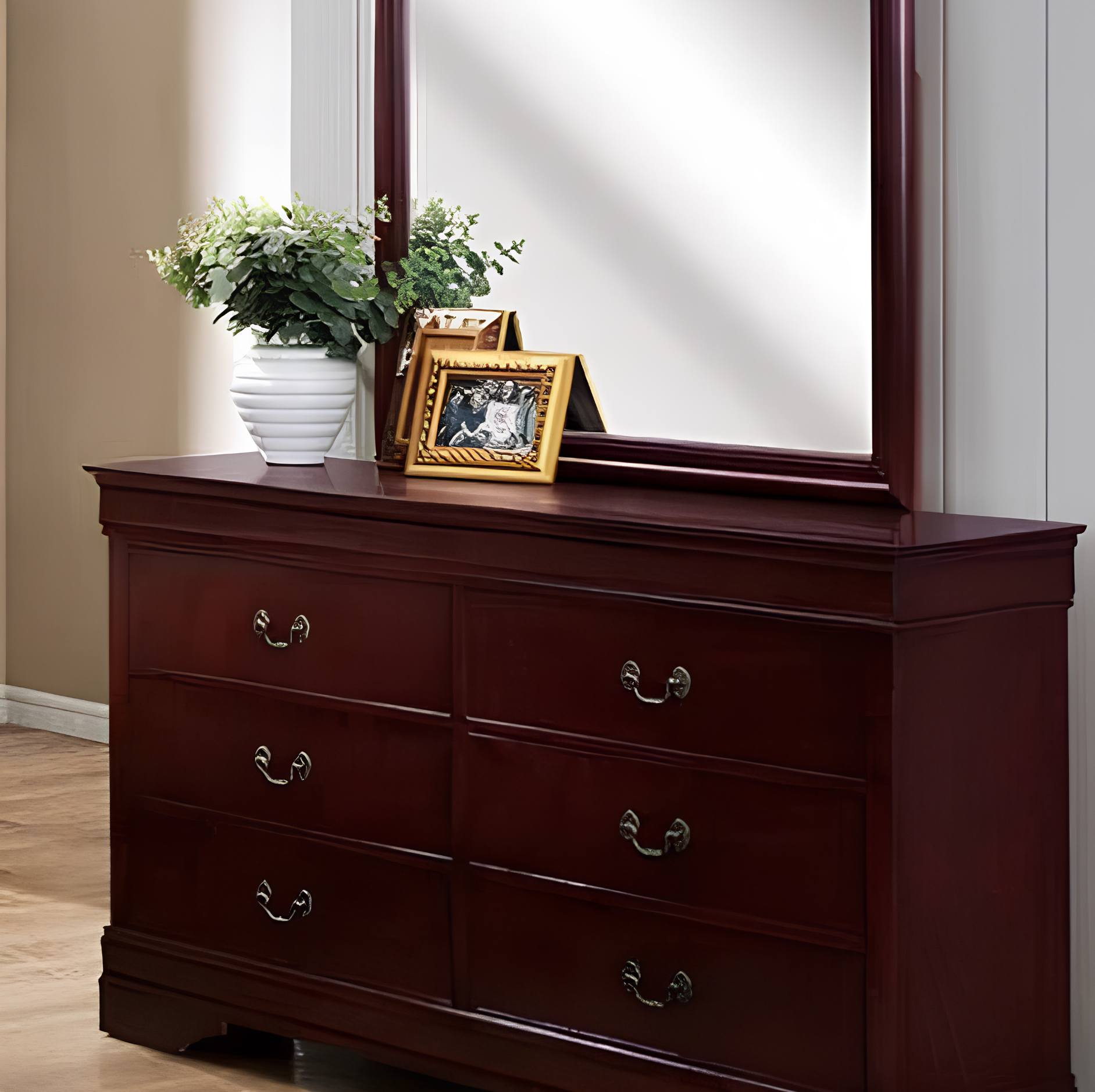 A dark reddish-brown wooden dresser with six drawers and silver metal handles, topped with a matching large mirror. A white planter with green leaves and two small picture frames sit on the dresser against a beige wall and white door frame.