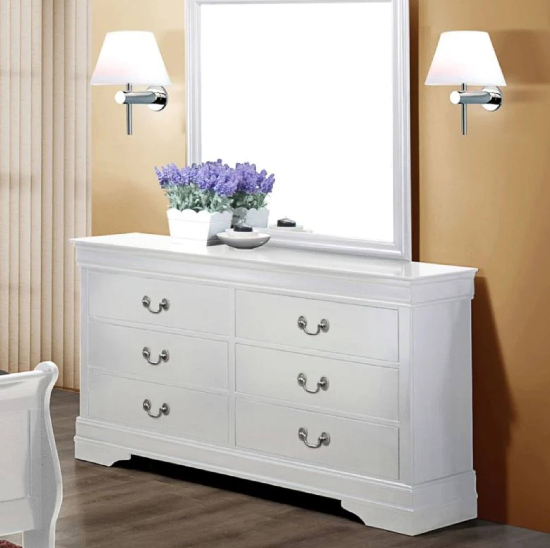 A white wooden dresser with six drawers and silver handles, styled in a bedroom setting with a large white mirror, two wall sconces, and a vase of purple flowers on top.