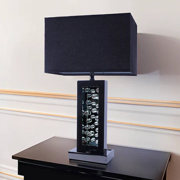 A modern table lamp with a large black rectangular fabric shade, a glossy black base featuring a vertical column of clear crystal-like spheres, and a silver-toned pedestal foot, shown on a dark side table against a cream wall with decorative molding.