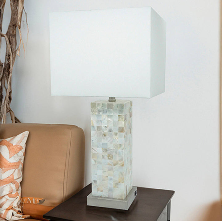 A table lamp with a large white rectangular shade and a mosaic shell-look base, shown on a dark side table next to a tan sofa in a home setting.