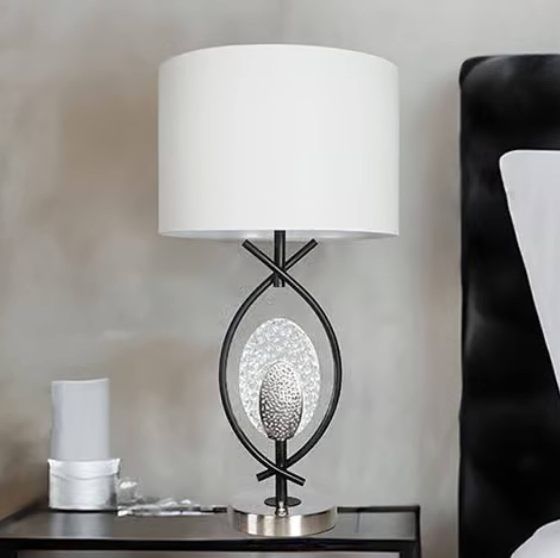 A modern table lamp with a white drum shade, a black metal frame, and a textured silver oval center, shown on a bedside table next to a black upholstered headboard.