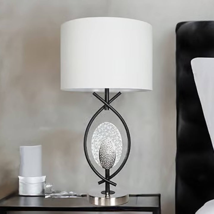 A modern table lamp with a white drum shade, a black metal frame, and a textured silver oval center, shown on a bedside table next to a black upholstered headboard.