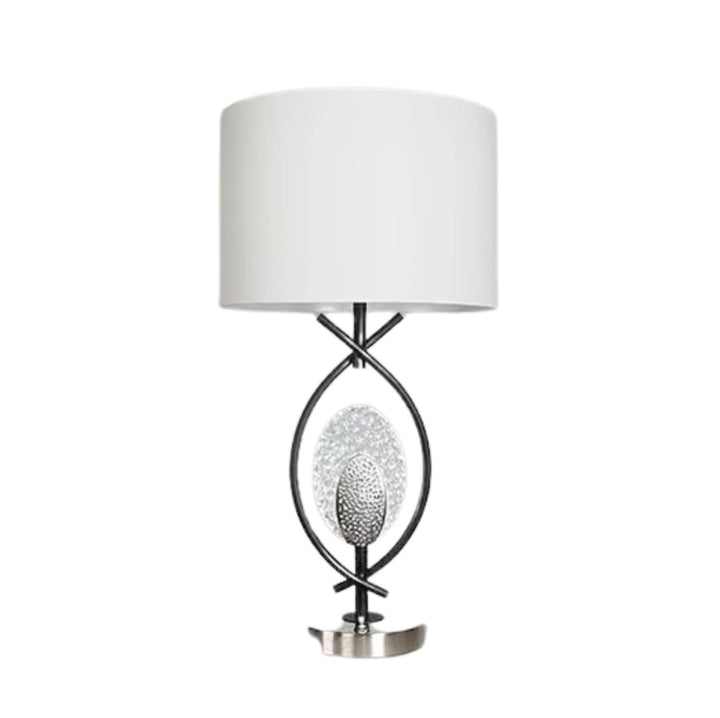 A modern table lamp with a white drum shade, a black metal frame, a textured silver oval center, and a polished silver base, shown against a plain white background.