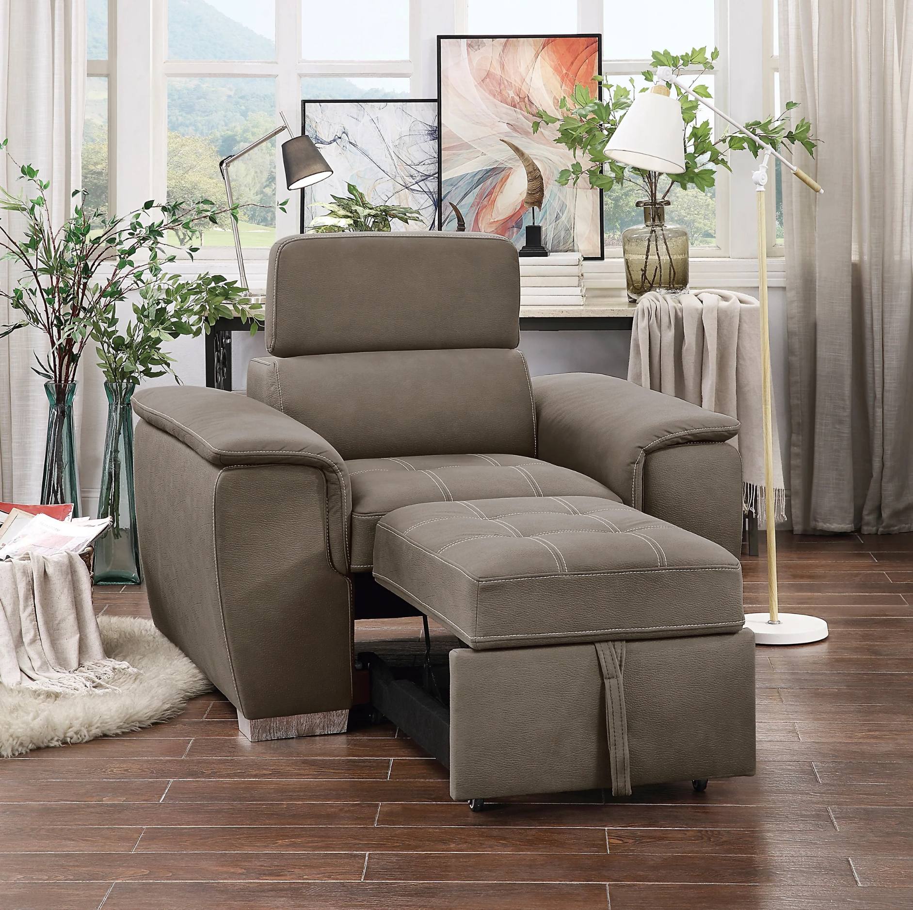 A taupe gray upholstered recliner chair with an attached matching ottoman, shown in a styled living room setting with wood floors, plants, and window decor.