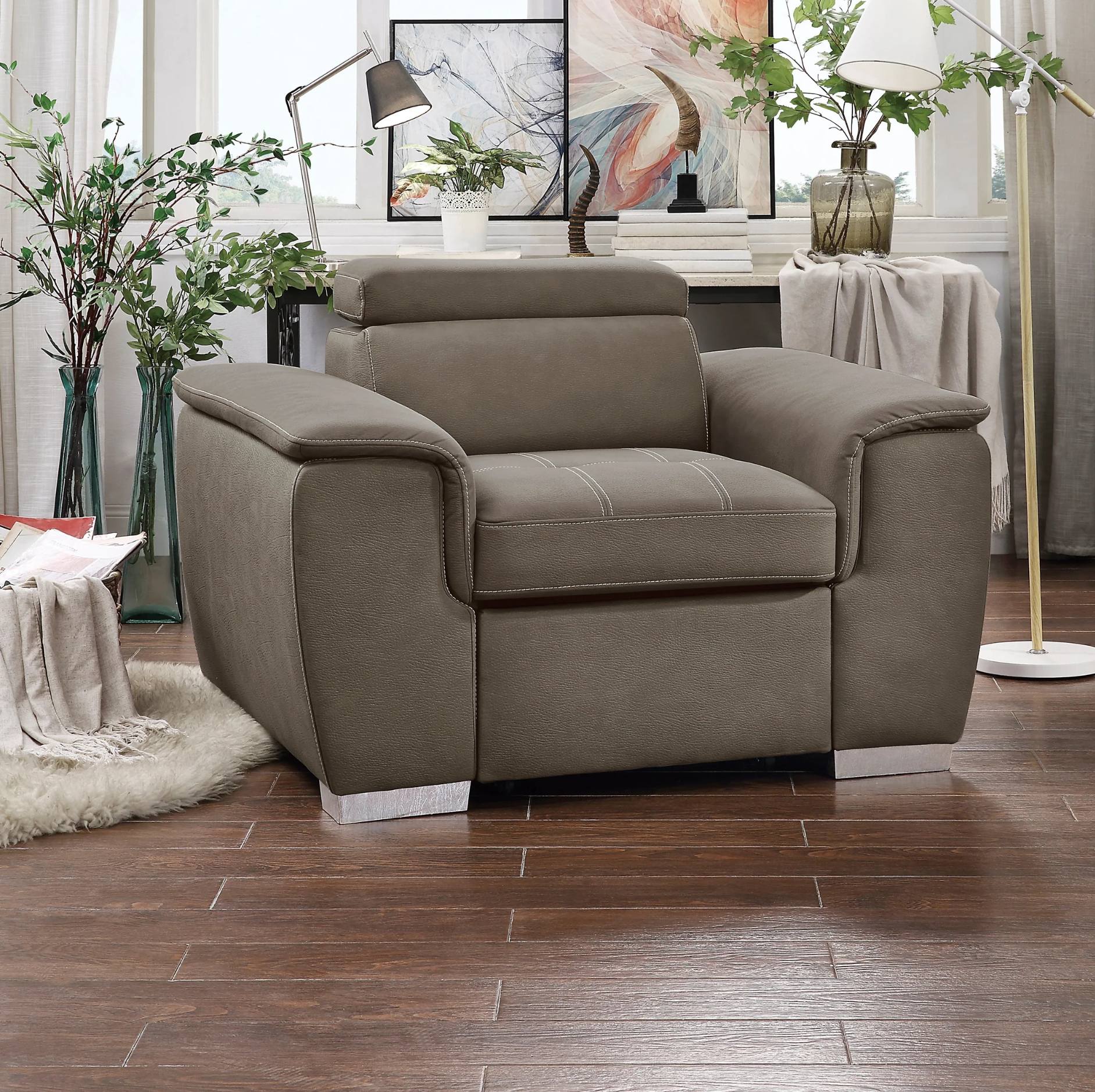 A taupe upholstered armchair with square arms and silver metal legs, shown in a styled living room setting with a wooden floor, decorative plants, and a lamp.