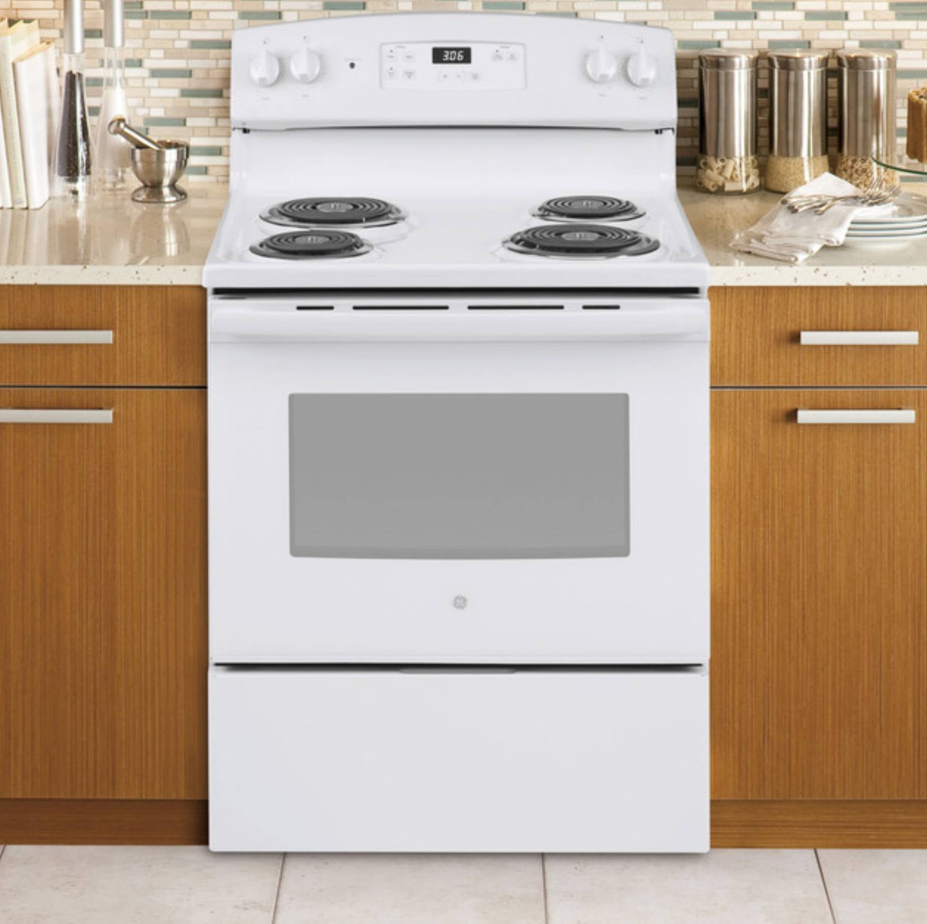 A white electric range with a glass oven door and four coil burners, set into a kitchen with wood cabinets and a tiled backsplash.