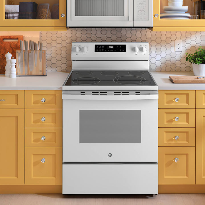 A white GE freestanding electric range with a black glass cooktop, set in a kitchen with yellow cabinets and a beige hexagonal tile backsplash.